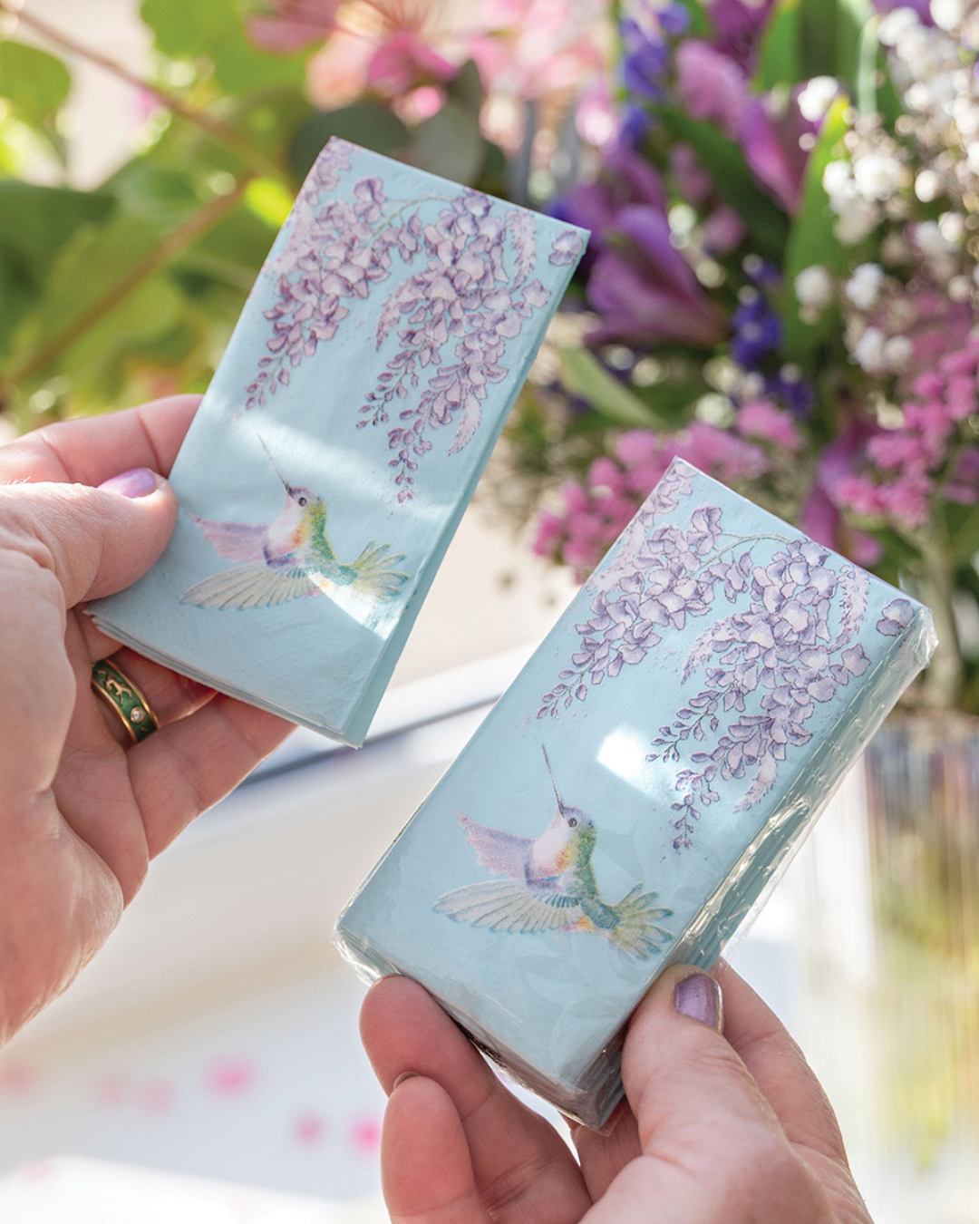 Eine Person hält in der linken Hand ein einzelnes türkisfarbenes Kolibri-Taschentuch mit violetten Glyzinien von Wrendale Designs und in der rechten Hand die geöffnete Packung, aus der es entnommen wurde; im Hintergrund sind unscharf bunte Blumen zu sehen.
