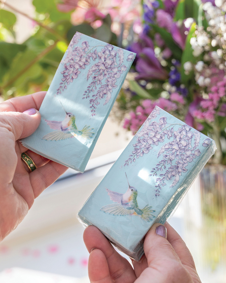 Eine Person hält in der linken Hand ein einzelnes türkisfarbenes Kolibri-Taschentuch mit violetten Glyzinien von Wrendale Designs und in der rechten Hand die geöffnete Packung, aus der es entnommen wurde; im Hintergrund sind unscharf bunte Blumen zu sehen.