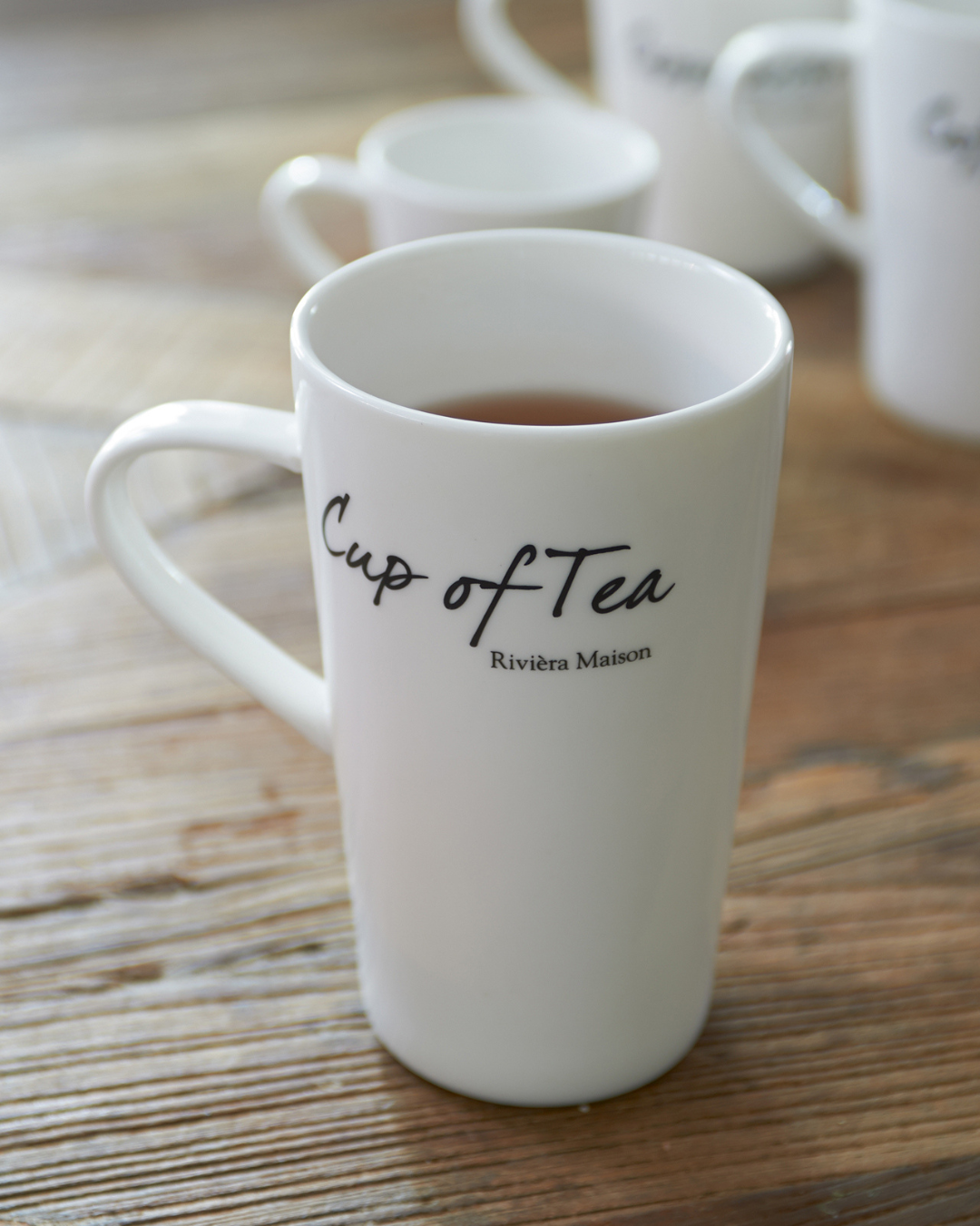 Classic Cup of Tea Tasse von Rivièra Maison, weiße Porzellantasse mit Schriftzug „Cup of Tea“, auf Holztisch stehend und mit dunklem Tee gefüllt.