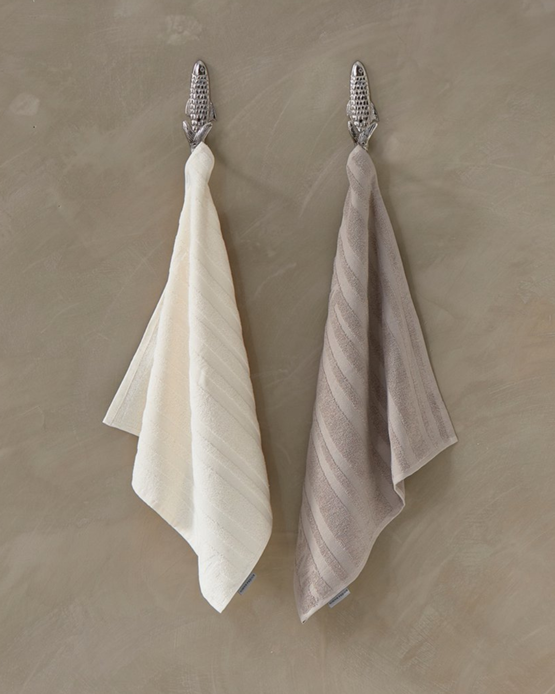 Zwei Ubuntu Küchenhandtücher von Rivièra Maison, in Beige und Weiß, dekorativ an Haken aufgehängt.