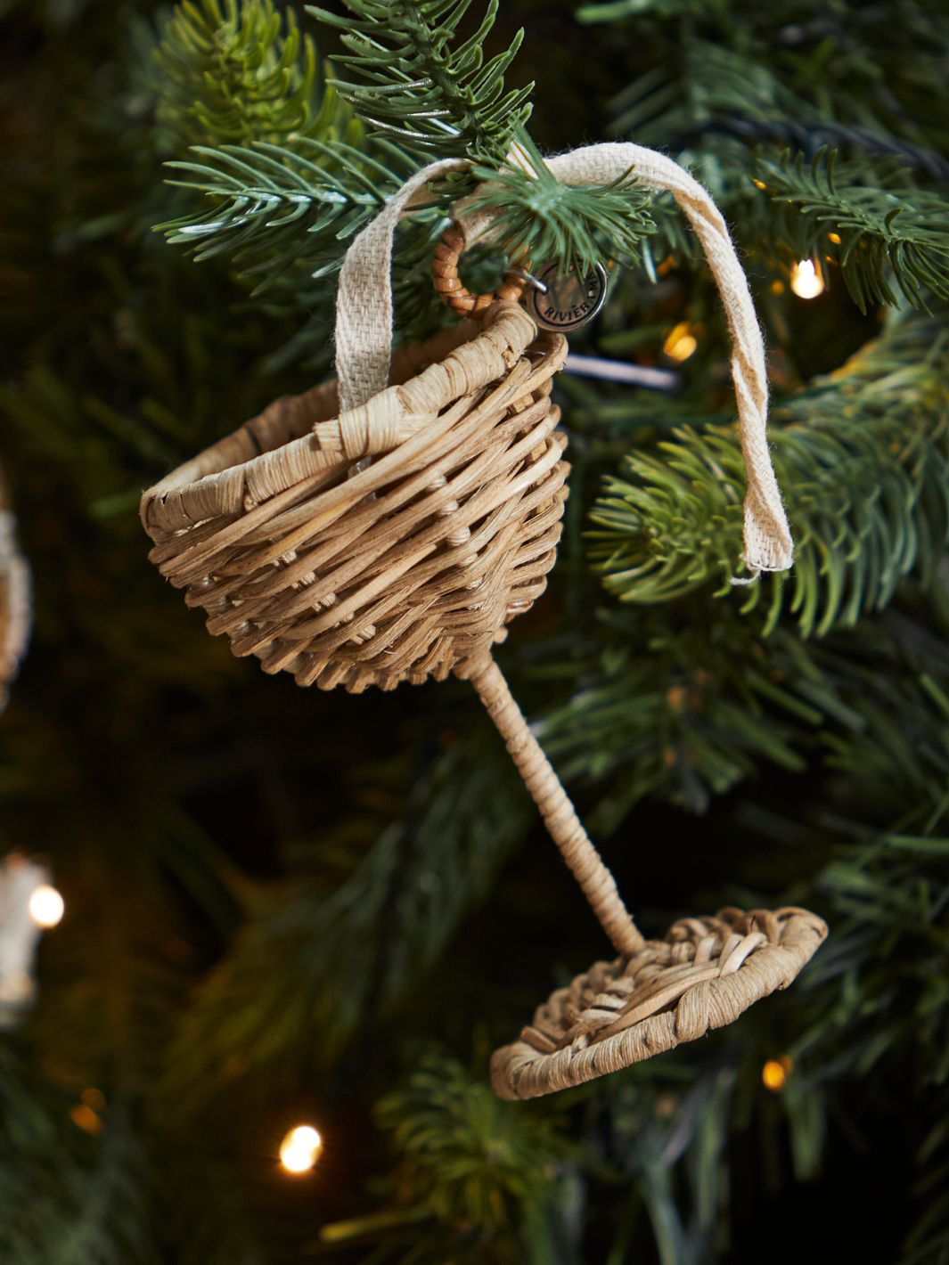 Rustic Rattan Champagnerglas Ornament von Rivièra Maison, aufgehängt an einem geschmückten Weihnachtsbaum mit Lichterkette.