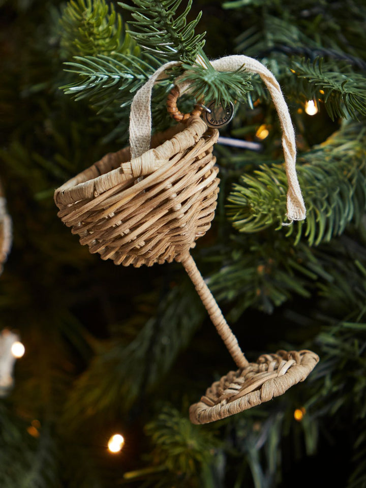 Rustic Rattan Champagnerglas Ornament von Rivièra Maison, aufgehängt an einem geschmückten Weihnachtsbaum mit Lichterkette.