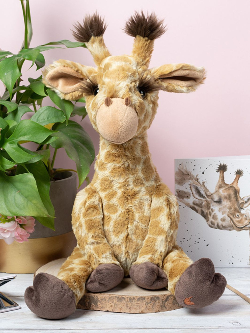 Wrendale Designs Kuscheltier Giraffe Camilla auf einer Baumscheibe neben einer Zimmerpflanze dekoriert.