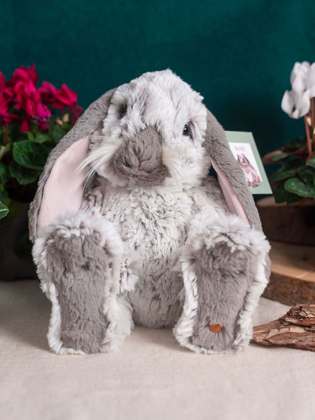 Der Wrendale Designs Kuscheltier Hase mit dem Namen Rosie sitzt vergnügt in einer Innenszene mit Alpenveilchen und schaut freundlich in die Kamera.