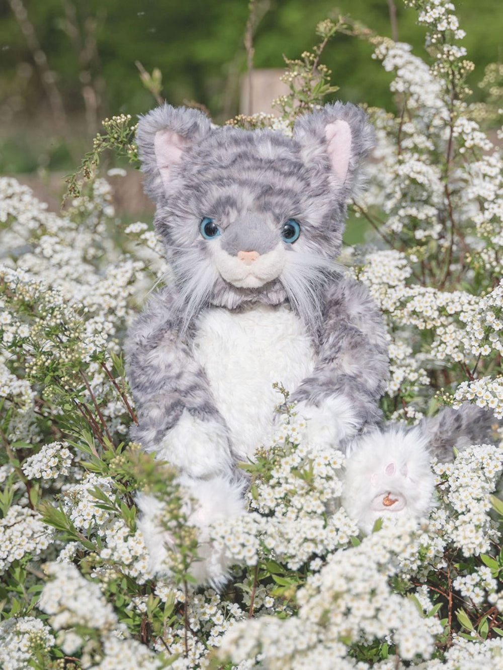 Das Wrendale Designs Kuscheltier Katze Esmeralda in den Farben grau und weiß sitzt inmitten von Blumen.