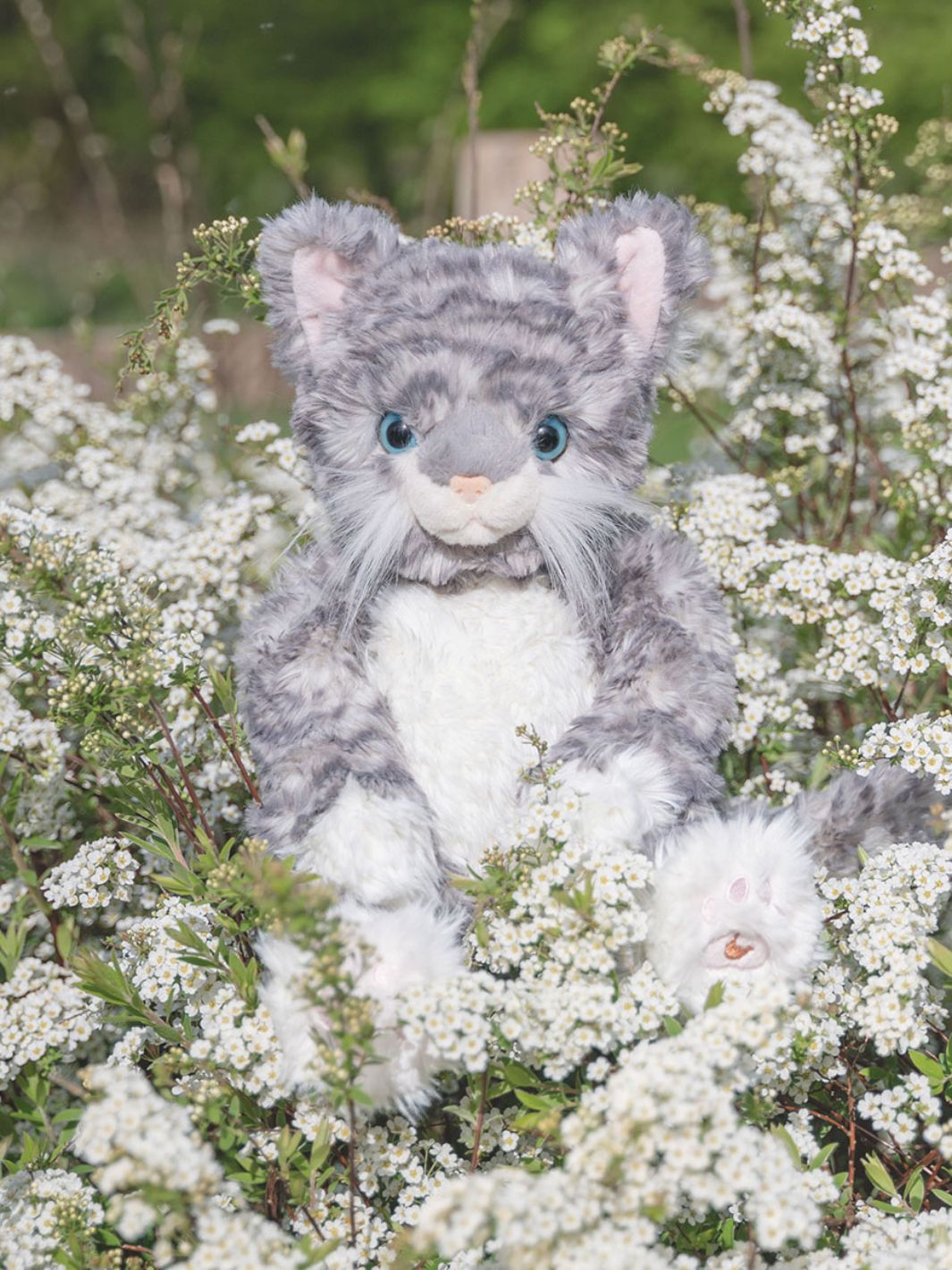 Das Wrendale Designs Kuscheltier Katze Esmeralda in den Farben grau und weiß sitzt inmitten von Blumen.