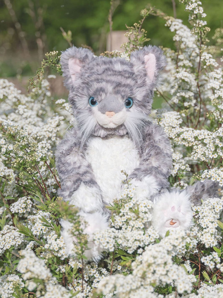 Das Wrendale Designs Kuscheltier Katze Esmeralda in den Farben grau und weiß sitzt inmitten von Blumen.