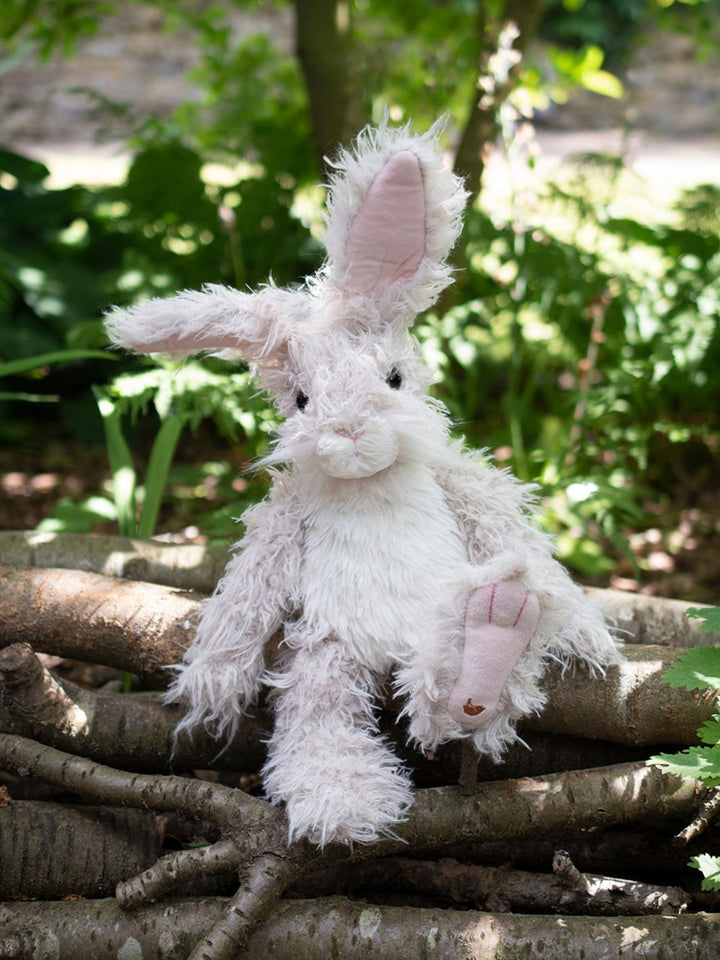 Der Wrendale Designs Kuscheltier Hase Rowan in den Farben grau und weiß sitzt im Wald.