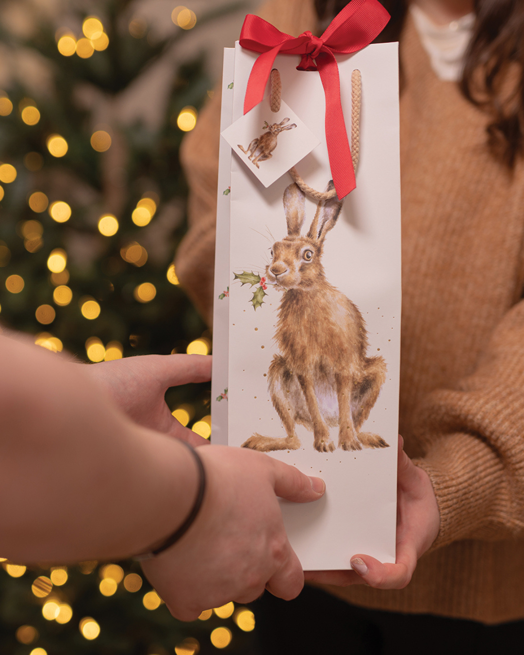 Person reicht Flaschen-Geschenktasche „Golden Hour“ mit roter Schleife vor einem Weihnachtsbaum einer anderen Person.