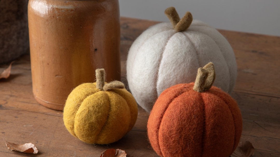 Drei Kürbisse aus Filz von Gry & Sif in den Farben gelb, orange und weiß und in unterschiedlichen Größen sind auf einem Holztisch neben einer braunen Vase dekoriert.