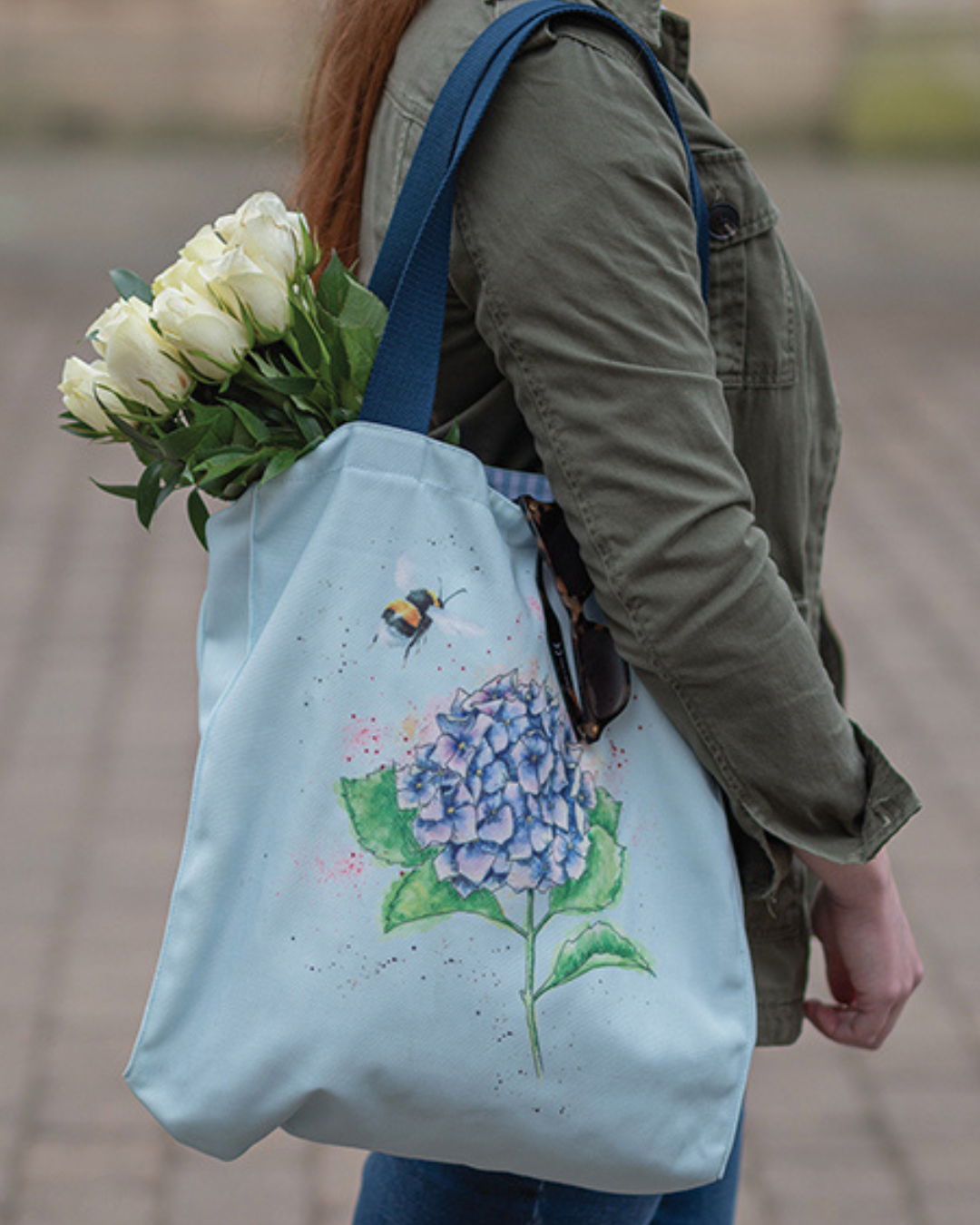 Hellblaue Hortensien Einkaufstasche von Wrendale Designs getragen von einer Frau. Das Motiv der Tasche ist eine lila Hortensie und eine Biene welche sich ihr nähert. Der Griff ist dunkelblau. Es hängen weiße Tulpen und eine Sonnenbrille aus der Tasche.