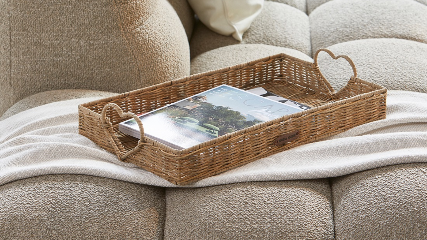 Ein großes Tablett mit Herzförmigen Griffen aus Rattan von Rivièra Maison ist auf einer hellen Couch dekoriert.