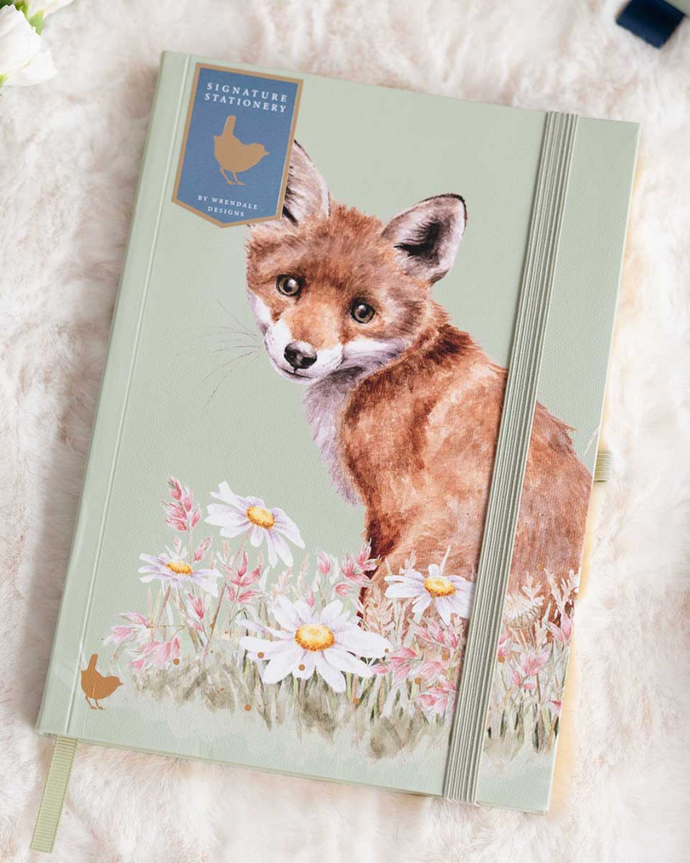 A5-Notizbuch „Fuchs“ aus der Signature Stationery Kollektion von Wrendale Designs. Motiv von Hannah Dale mit jungem Fuchs zwischen Margeriten, grünem Gummiband und goldfarbenem Seitenschnitt auf einer weißen Decke.