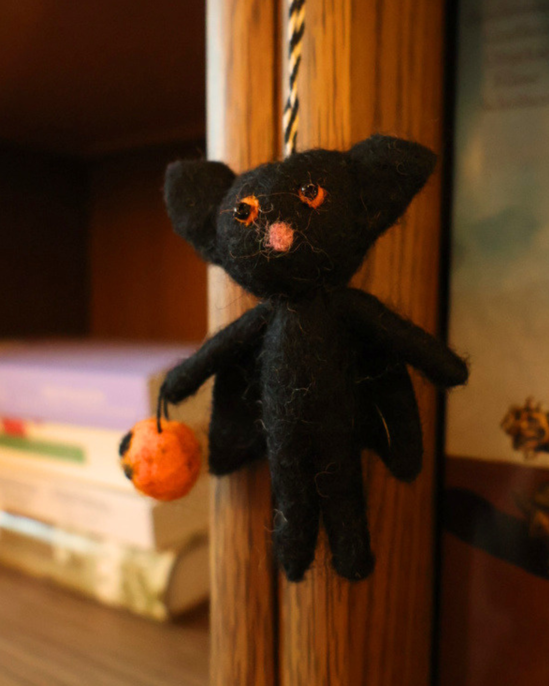 Dekorierter "Fledermaus" Filzanhänger von Grace Gloria hängt an Bücherregal. Zu sehen ist eine Fledermaus mit orangenen Augen und einem orangenem Körbchen.