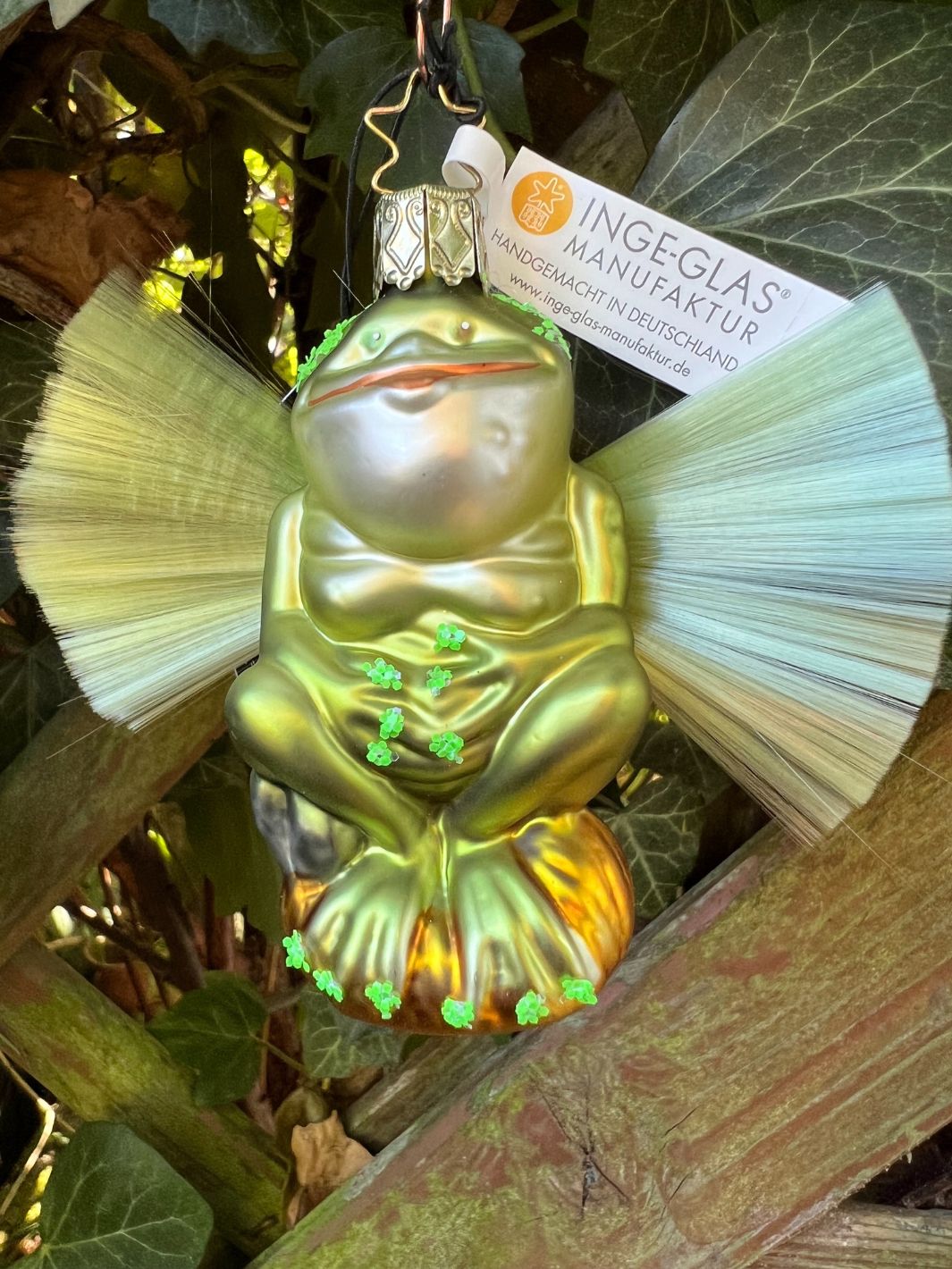 Das handgefertigte Frosch Ornament "Ariwur" von Inge Glas ist in farbenprächtigen Grün- ,Gelb-, Silber- und Goldtönen bemalt. Er sitzt buddha-ähnlich auf einem Stein. An seinem Rücken formen Kunststofffasern Flügel.