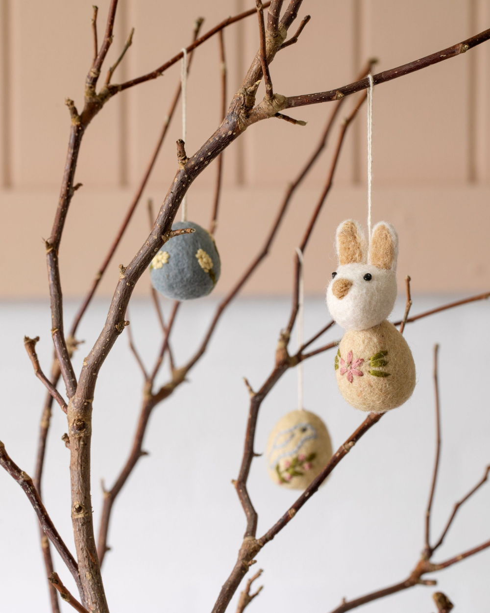 Filz-Osterei mit Blumen-Stickerei hängt an einem Naturast neben einem kleinen Hasen-Filzanhänger in warmer Wohnumgebung.
