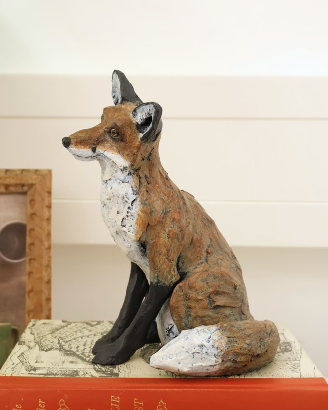 Fuchs Statue "Autumn" von Wrendale Designs auf Büchern sitzend. Im Hintergrund ist eine weiße Wand und ein Bild zu sehen.