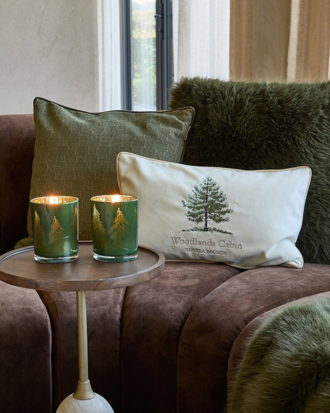 Auf einer braunen Couch sind die Kissen RM Monogram in grün , Forges und Bromont dekoriert. Dazu passend stehen davor auf einem kleinen Tisch zwei Windlichter mit Tannenbäumen drauf. Seitlich ist noch die dunkelgrüne Bromont Decke zu sehe.