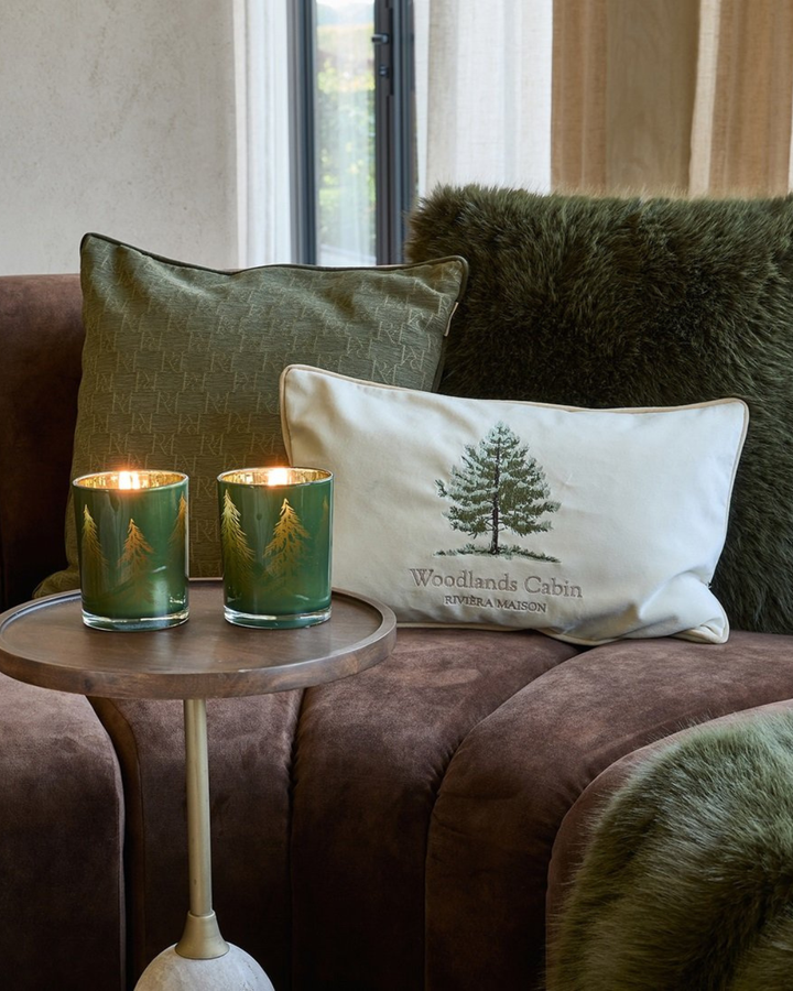 Auf einer braunen Couch sind die Kissen RM Monogram in grün , Forges und Bromont dekoriert. Dazu passend stehen davor auf einem kleinen Tisch zwei Windlichter mit Tannenbäumen drauf. Seitlich ist noch die dunkelgrüne Bromont Decke zu sehe.