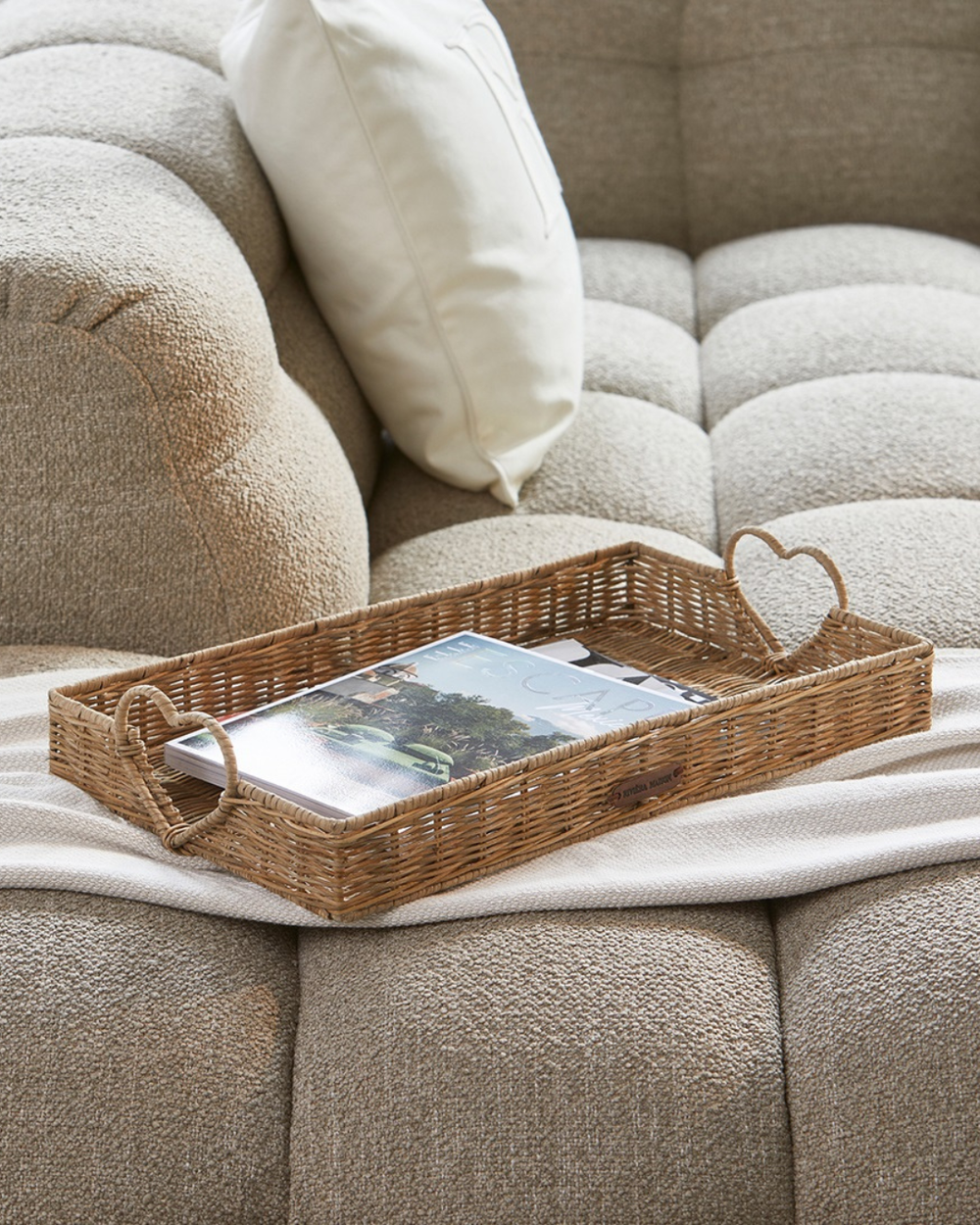 Rustic Rattan Tablett „Happy Heart Tray“ von Rivièra Maison mit Zeitschriften auf einer hellen Couch platziert.