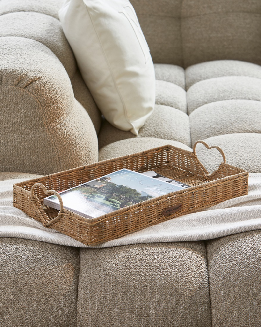 Rustic Rattan Tablett „Happy Heart Tray“ von Rivièra Maison mit Zeitschriften auf einer hellen Couch platziert.