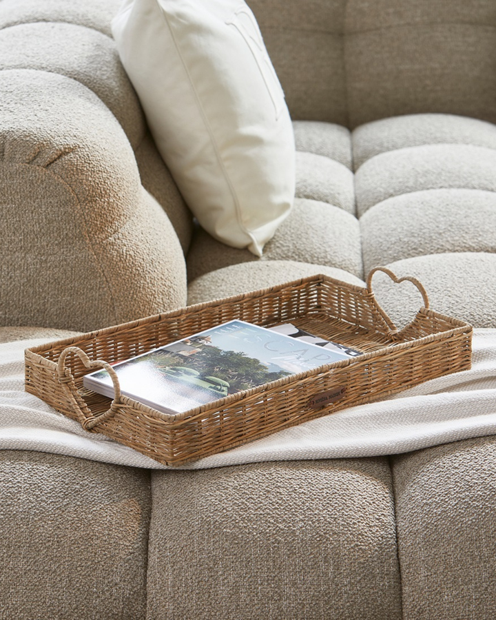 Rustic Rattan Tablett „Happy Heart Tray“ von Rivièra Maison mit Zeitschriften auf einer hellen Couch platziert.