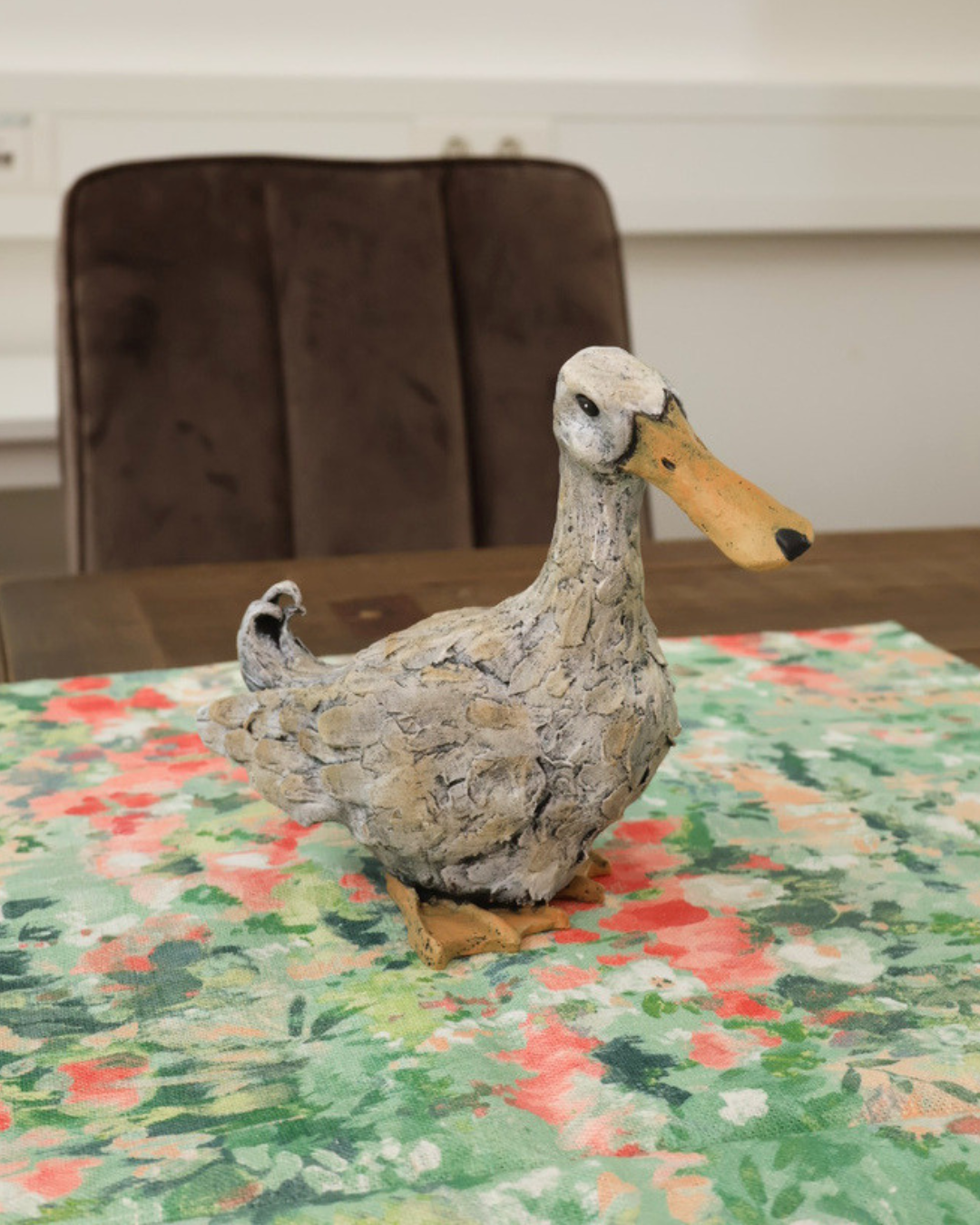 Enten Statue "Lily" auf dekorierten Holztisch. Auf Dem Tisch liegt ein Tuch mit Mustern. Die Ente schaut nach rechts unten.