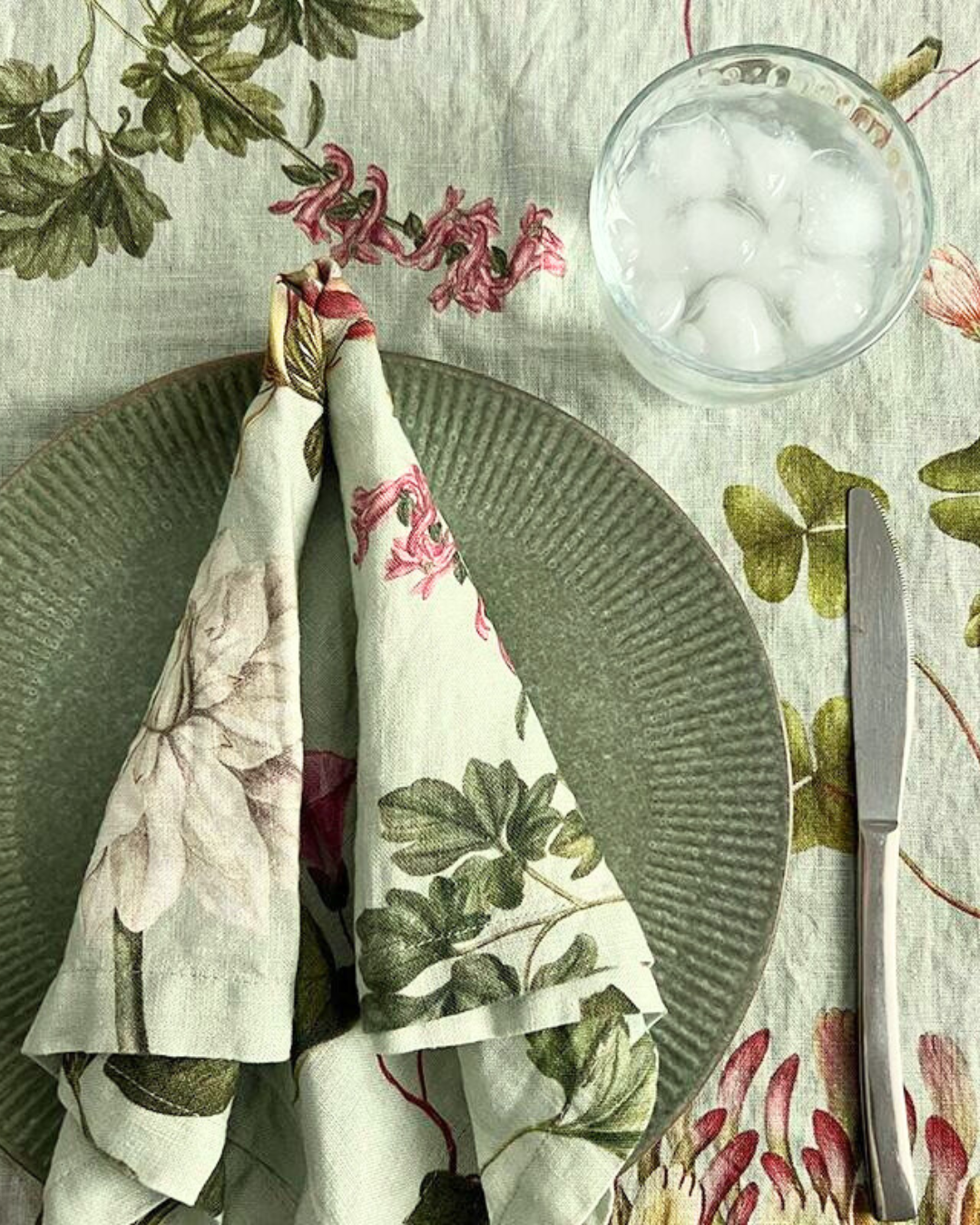 Leinenserviette "Flora Danica Atlas" von Koustrup & Co., gefaltet auf Tisch mit Besteck, Glas und botanischen Motiven auf Tischdecke.