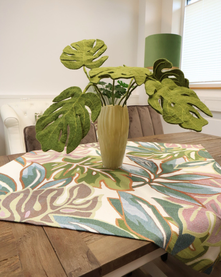 Zimmerpflanzen Mitteldecke Selva von Sander Table + Home auf einem Holztisch mit Vase. Die mehrfarbigen Monsterblätter sind sich gegenseitig überlappend dargestellt. Blaue, grüne und roséfarbene Blätter mit sich farblich absetzender Blattumrandung sind in den Stoff der Mitteldecke gewoben.