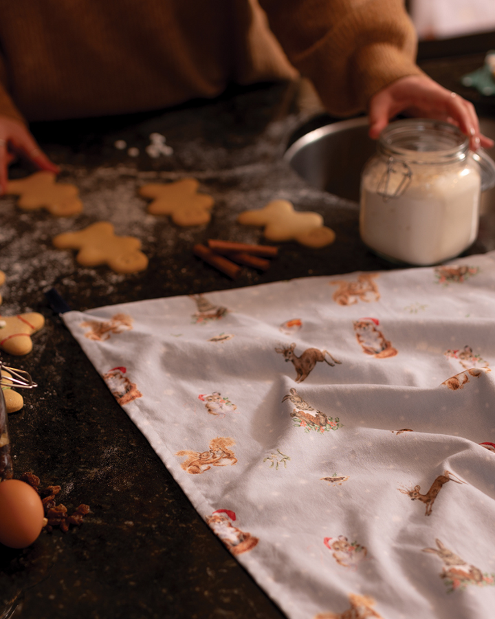 Ein weihnachtliches Geschirrtuch mit Waldtiermotiven liegt auf einer dunklen Küchenarbeitsfläche. Daneben stehen ein Glas mit Mehl, Zimtstangen und ausgestochene Plätzchen in Form von Lebkuchenmännern.