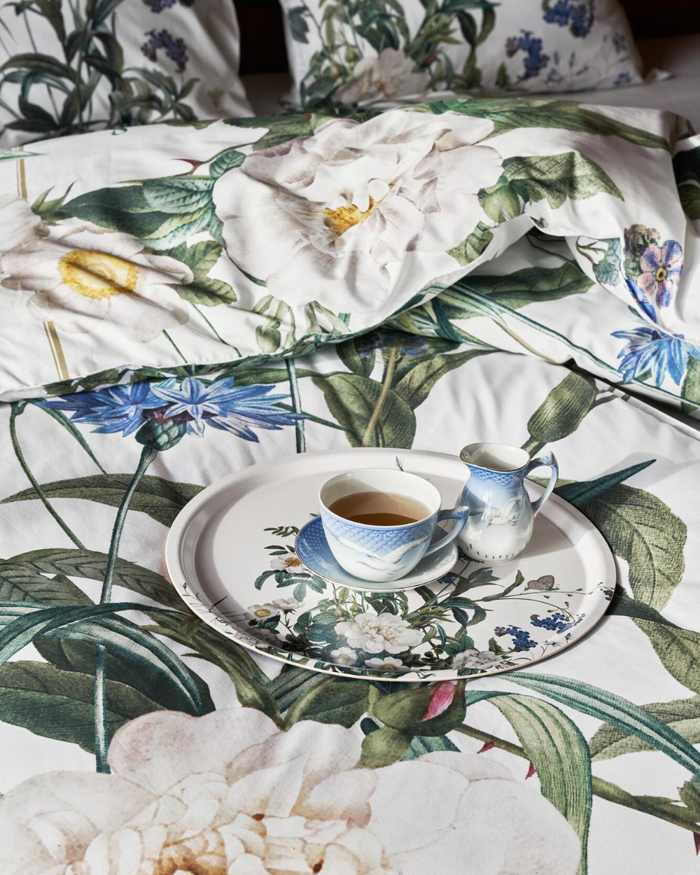 Detailaufnahme der Bettwäsche „Blue Flower Garden“ mit Teeservice auf dem Bett, große weiße Rosen und blaue Glockenblumen im Vordergrund.