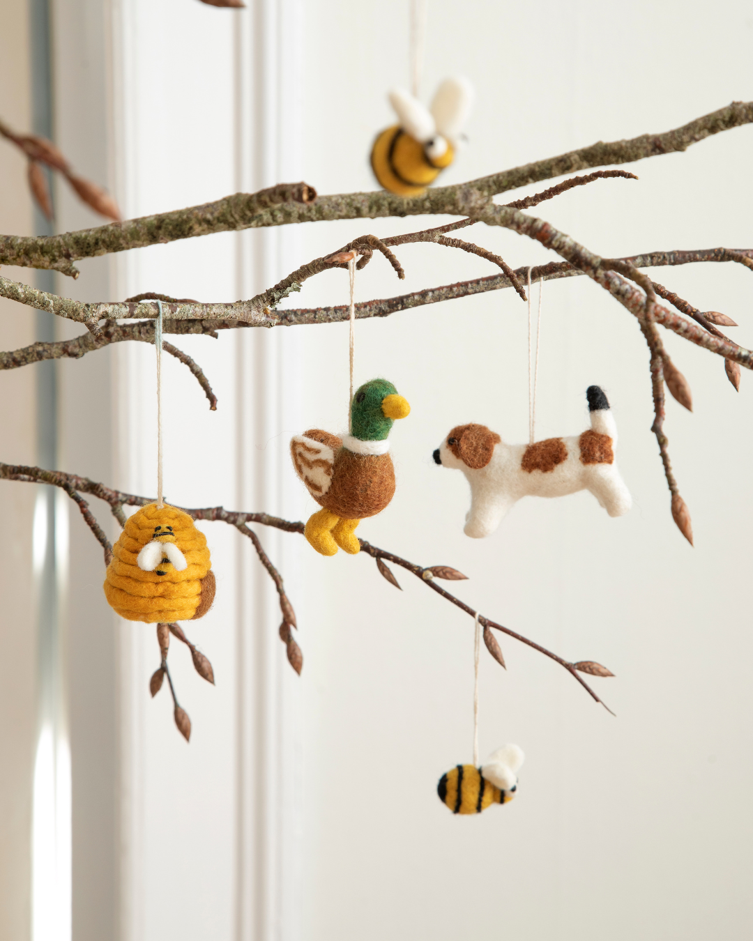 Deko-Ast vor weißer Wand mit verschiedenen Filzanhängern von Gry & Sif – Erpel, Bienenstock, Bienen und Hund bereichern diese harmonische Dekoration.