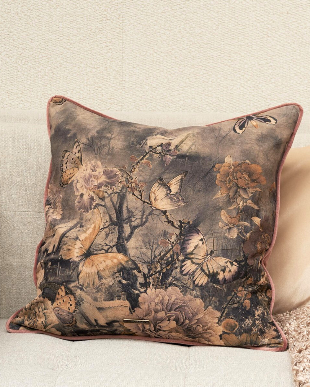 Das Kissen La Belle Epoque von Rivièra Maison ist auf einer beigen Couch dekoriert.
