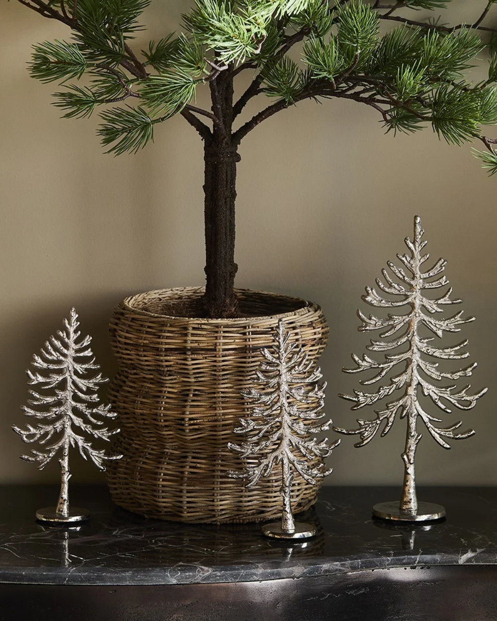3 dekorierte "Alpina Christmas Tree" Statuen von Riviera Maison. Die Statuen liegen auf einem Schwarzen Tisch vor einem mini Nadelbaum in einem Blumentopf.