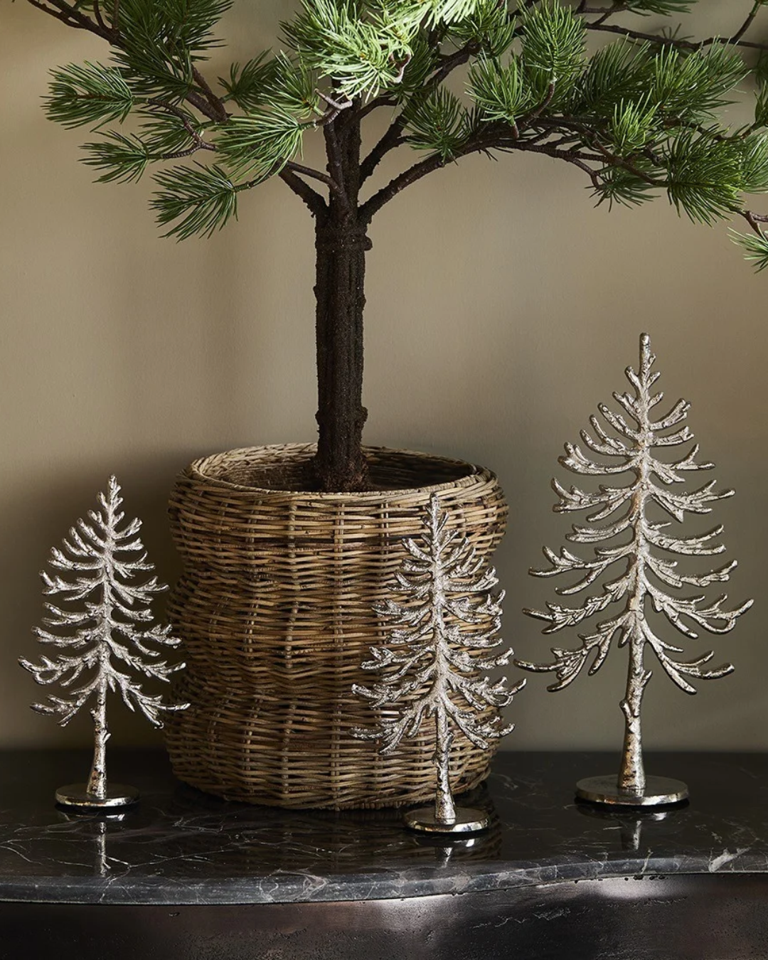 3 dekorierte "Alpina Christmas Tree" Statuen von Riviera Maison. Die Statuen liegen auf einem Schwarzen Tisch vor einem mini Nadelbaum in einem Blumentopf.