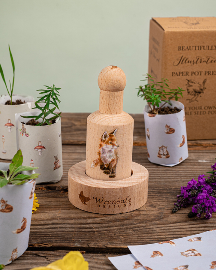 Dekorative Szene mit Anzuchttopf-Presse von Wrendale Designs mit Fuchs-Motiv auf Holztisch, umgeben von selbstgemachten Papier-Anzuchttöpfen mit kleinen Pflanzen und Saatgutpapier.