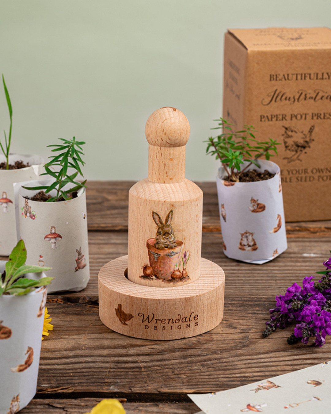 Dekorative Szene mit Anzuchttopf-Presse von Wrendale Designs mit Hasen-Motiv auf rustikalem Holztisch, mehrere bepflanzte Papier-Anzuchttöpfe im Hintergrund, natürliche Gartenatmosphäre.