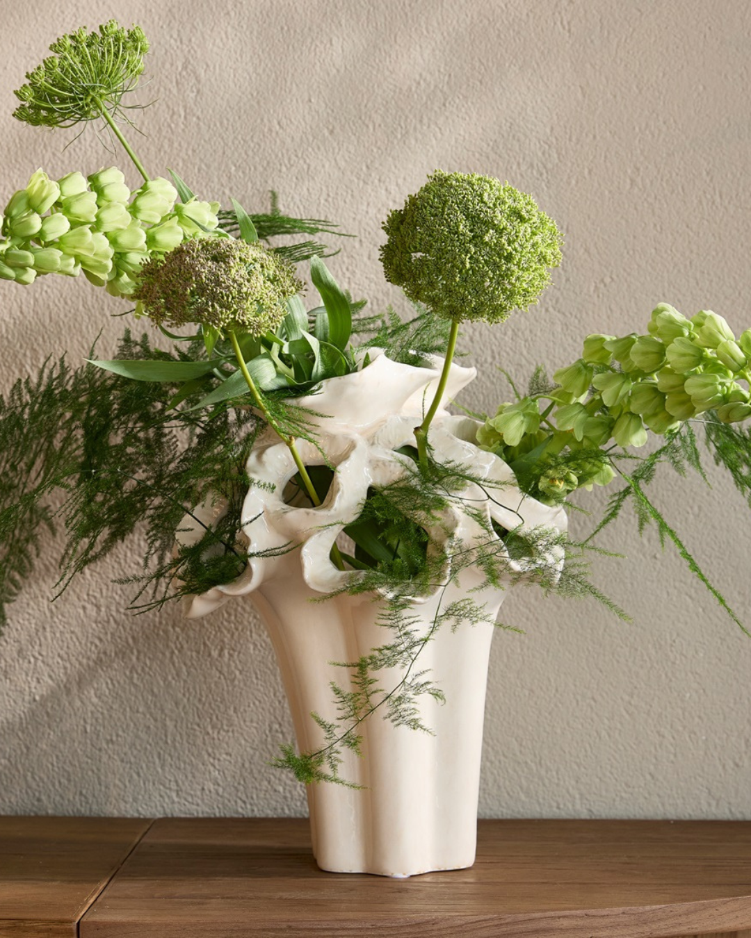 Dekorierte Vase "Atelier Flora" in Größe M von Riviera Maison. Die weiße kunstvolle Vase steht auf einem etwas dunkleren Holztisch. Die Vase ist gefüllt mit Blumen/Pflanzen.