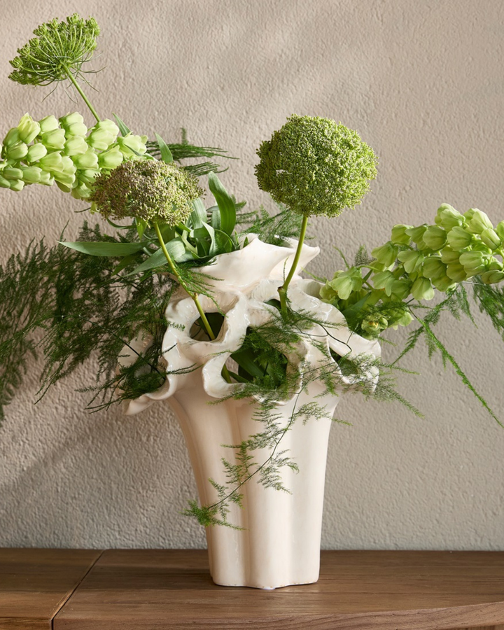 Dekorierte Vase "Atelier Flora" in Größe M von Riviera Maison. Die weiße kunstvolle Vase steht auf einem etwas dunkleren Holztisch. Die Vase ist gefüllt mit Blumen/Pflanzen.