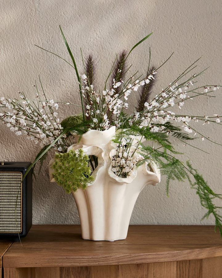 Dekorierte Vase "Atelier Flora" in Größe S von Riviera Maison. Die weiße kunstvolle Vase steht auf einem etwas dunkleren Holztisch und ist mit Blumen/Pflanzen beschmückt.