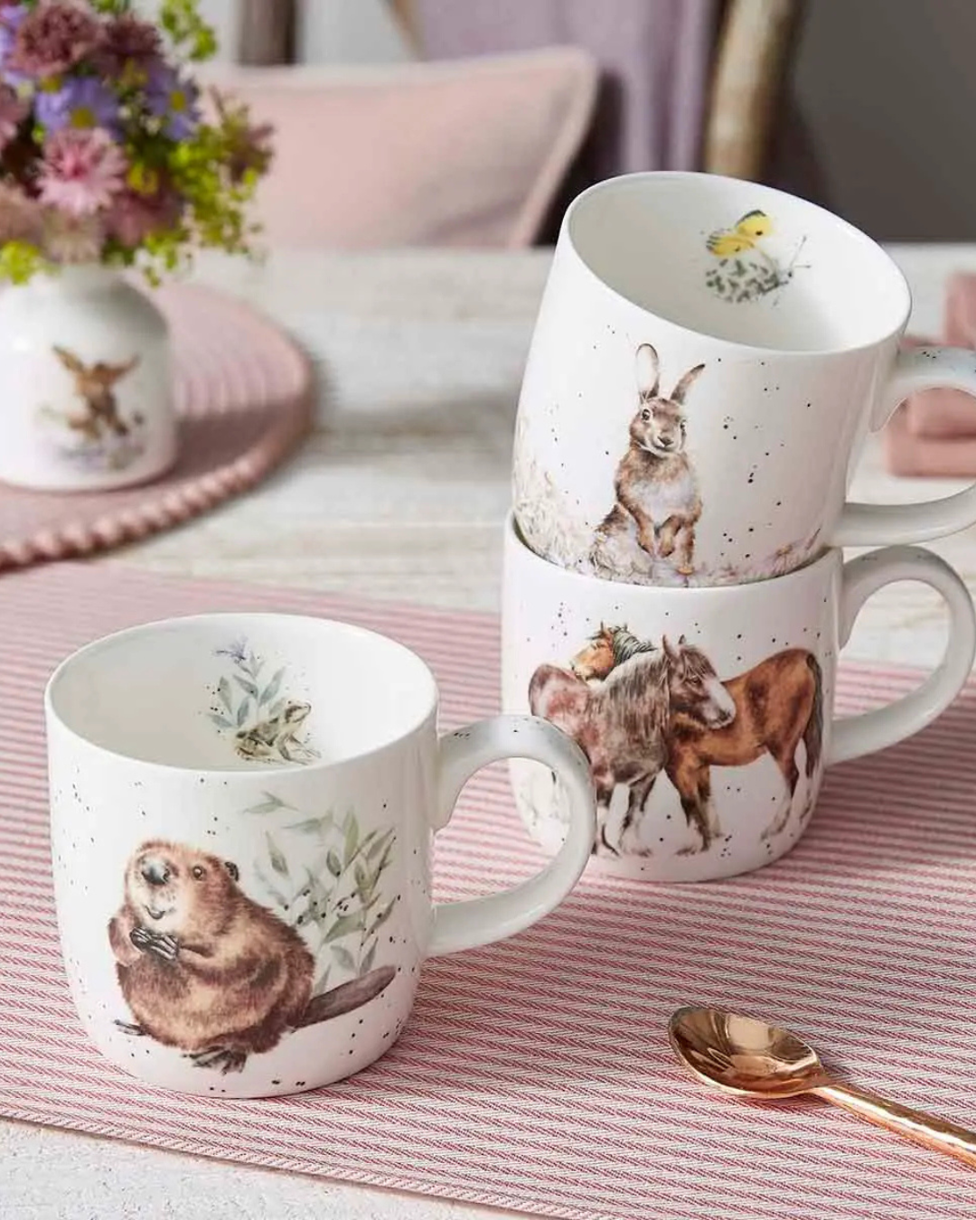 Es sind 3 Tassen von Wrendale Designs auf einem größtenteils pink gedeckten hellen Holztisch zu sehen. Die Tassen haben 3 verschiedene Motive: ein Biber, ein Hase und 2 Pferde.