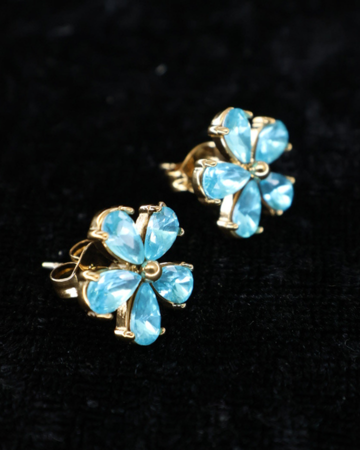 Blumenohrringe von "Dottilove" in hellem blau mit goldenen Akzenten welche auf einer Schwarzen leicht glitzernden Decke liegen.
