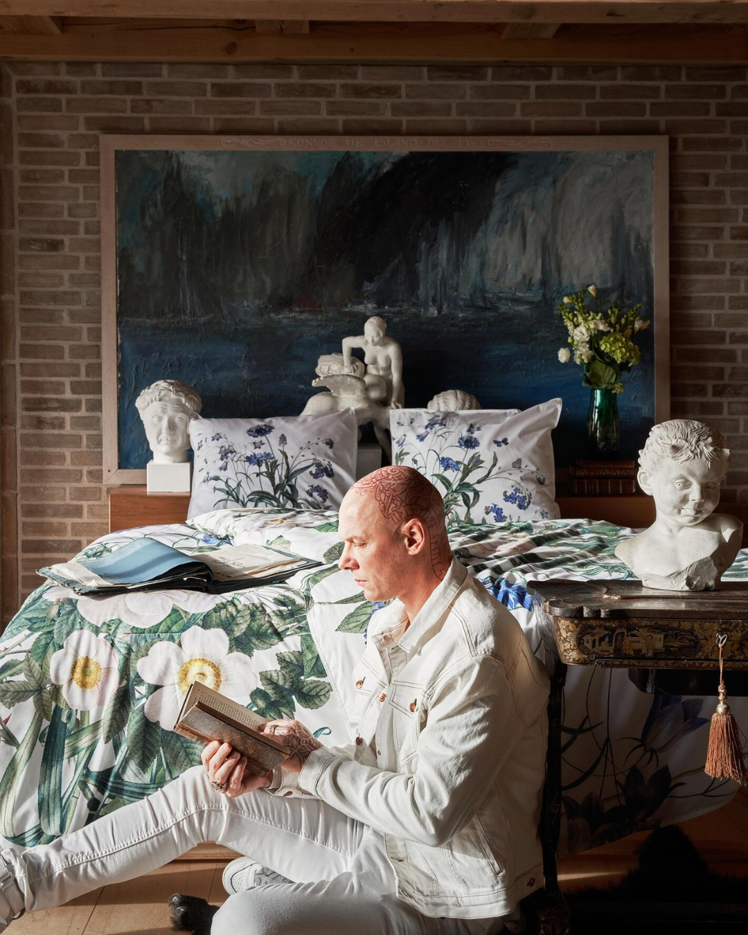 Person sitzt auf einem Bett mit der Bettwäsche „Blue Flower Garden“, umgeben von Büsten und Kunstwerken, gemütliche Leseatmosphäre.