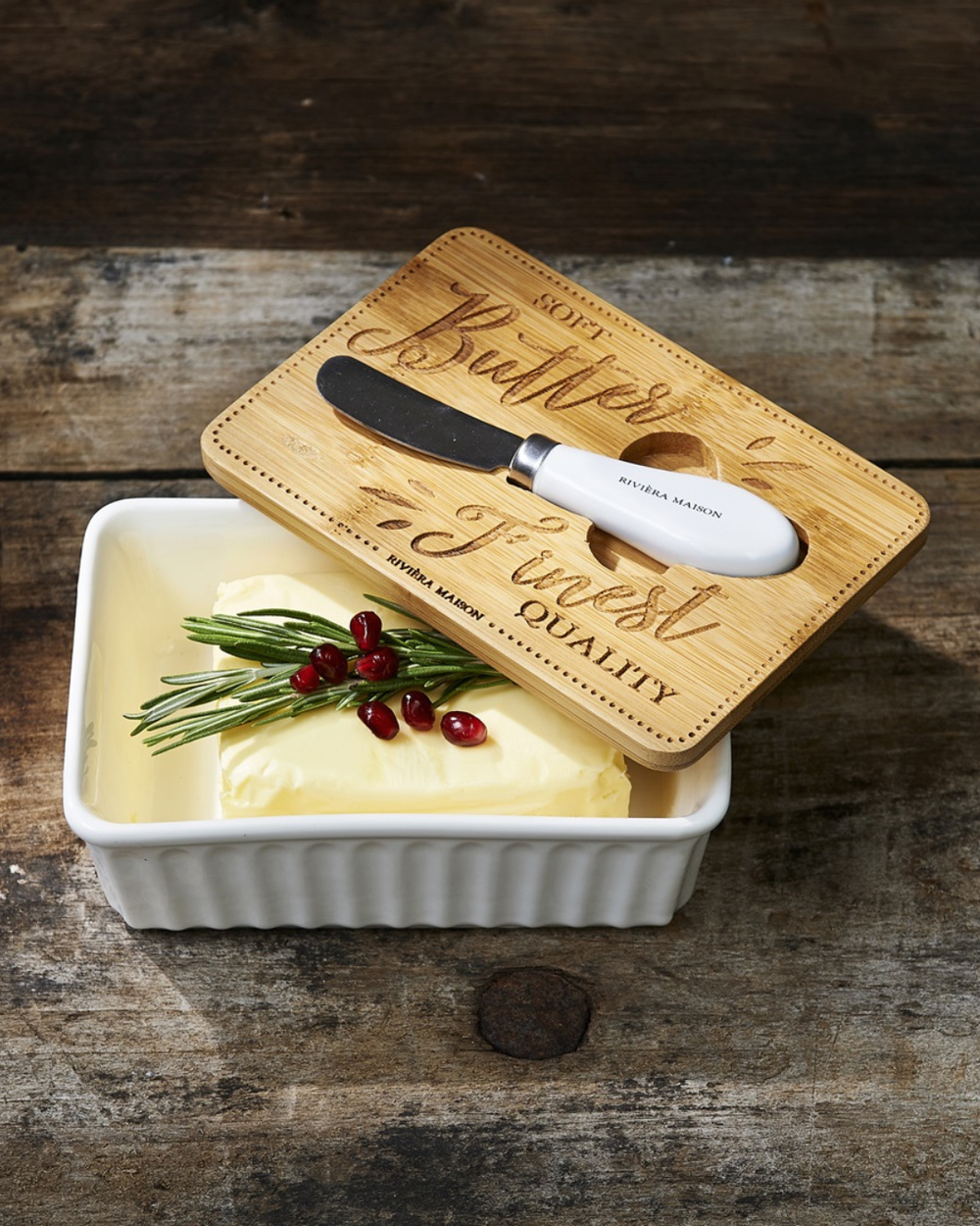 Butterdose „Finest Quality Butter Dish“ von Rivièra Maison mit Butter, dekoriert mit Rosmarin und Granatapfel und kleinem Messer auf dunklem Tisch.