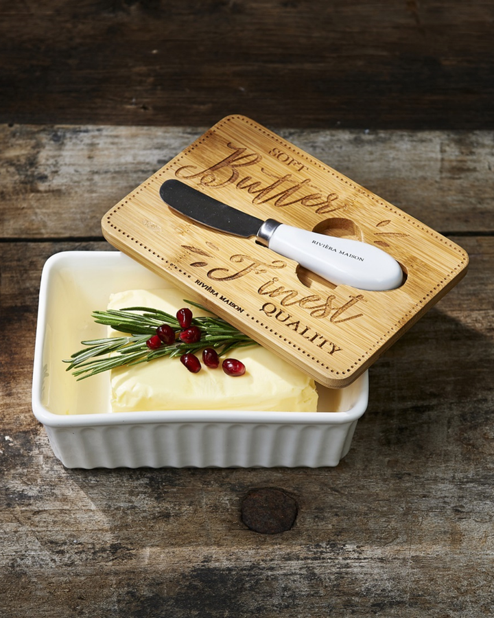 Butterdose „Finest Quality Butter Dish“ von Rivièra Maison mit Butter, dekoriert mit Rosmarin und Granatapfel und kleinem Messer auf dunklem Tisch.