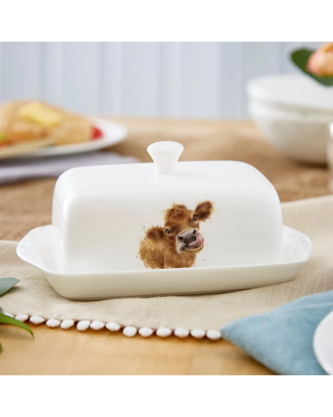 Kuh Butterdose "Royal Worchester" aus Porzellan von Wrendale Designs auf einem bedeckten Tisch. Das Motiv zeigt eine Kuh mit herausgestreckter Zunge. Im Hintergrund sind Teller zu sehen.