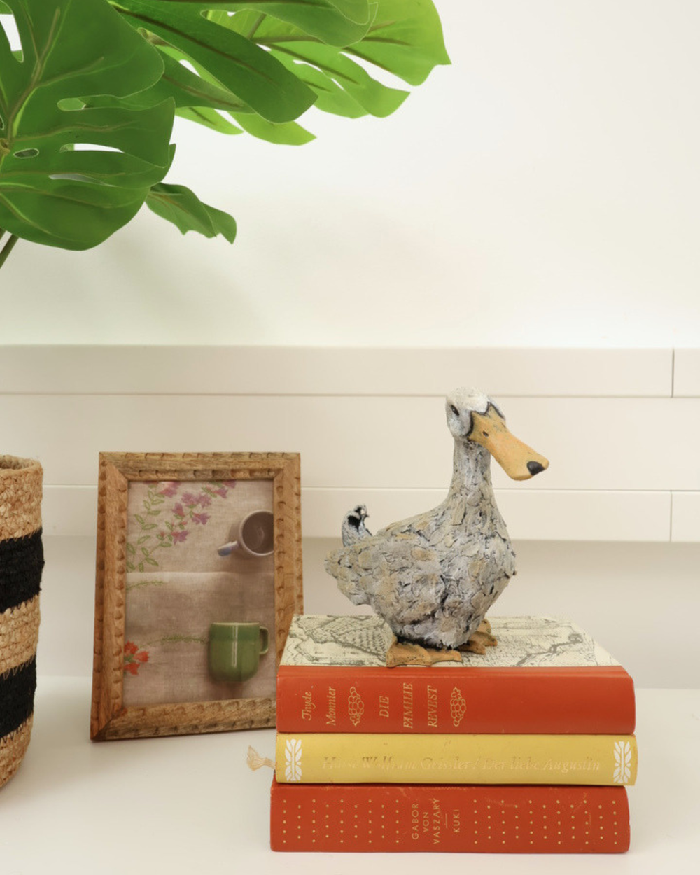 Enten Statur "Lily" auf dekorierten weißen Tisch. Die Statue steht auf 3 Büchern neben einem Bild und einer Zimmerpflanze.