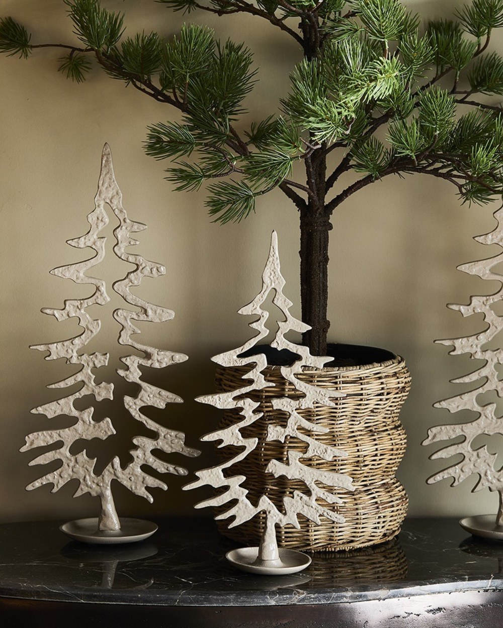 Dekorierten "Festive Christmas Tree Statuen" von Riviera Maison. zu sehen ist ein Schwarzer Tisch mit 3 Statuen in unterschiedlichen Größen vor einem mini Nadelbaum.