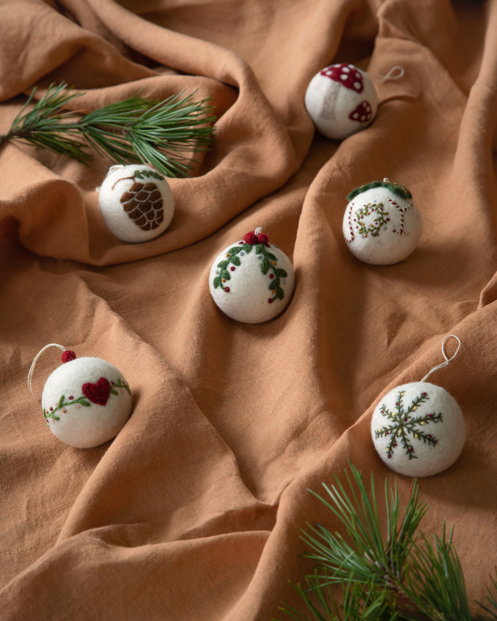Mehrere handgefertigte Weihnachtsbaumkugeln aus weißem Filz von Gry & Sif, liegend auf einem braunen Stoff mit Tannenzweigen dekoriert. Jede Kugel ist unterschiedlich bestickt, unter anderem mit Herz, Kranz, Tannenzapfen, Schneeflocke und Pilzmotiv.