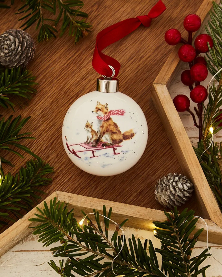 Dekorierte Weihnachtsbaumkugel „Schlittenfahrt der Füchse“ von Wrendale Designs auf einem Sterntablett aus Holz.